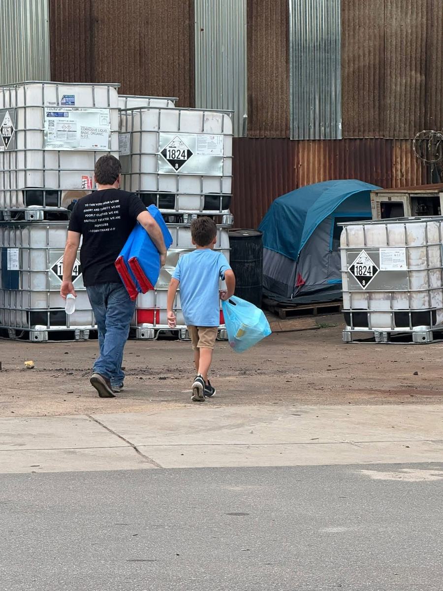 A man and a boy walk away from the camera carrying a sleeping mat, a water bottle and a bag of supplies toward a tent pitched behind a warehouse