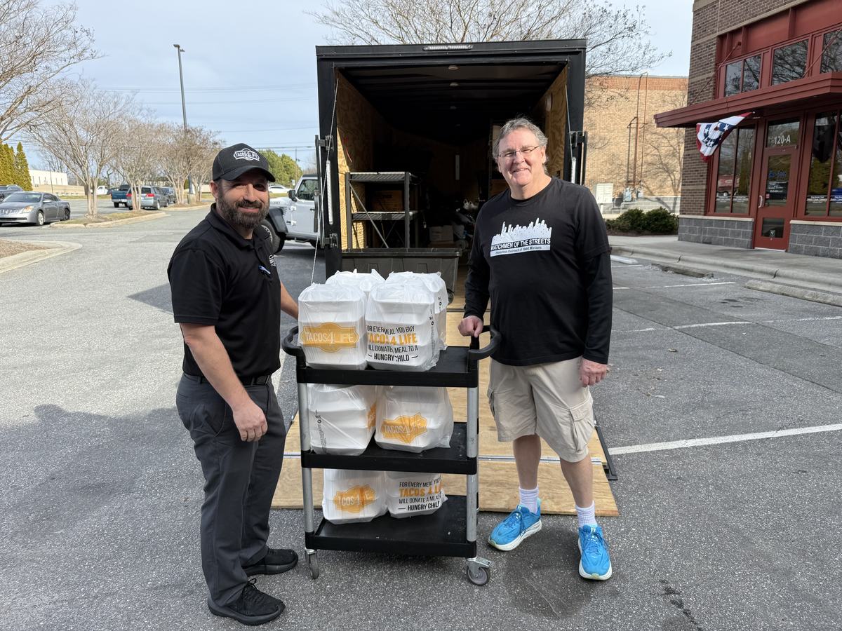 Volunteers loading racks of packed meals into a van outside a restaurant